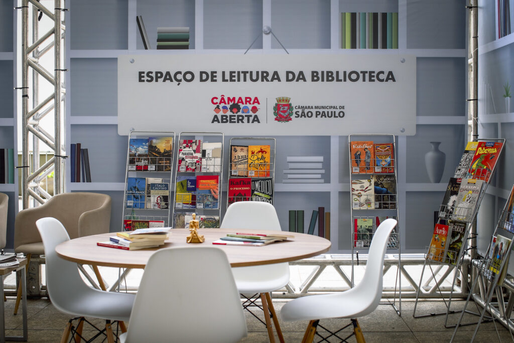 Espaço de Leitura da Biblioteca no Câmara Aberta, com mesas, cadeiras e livros em revisteiros