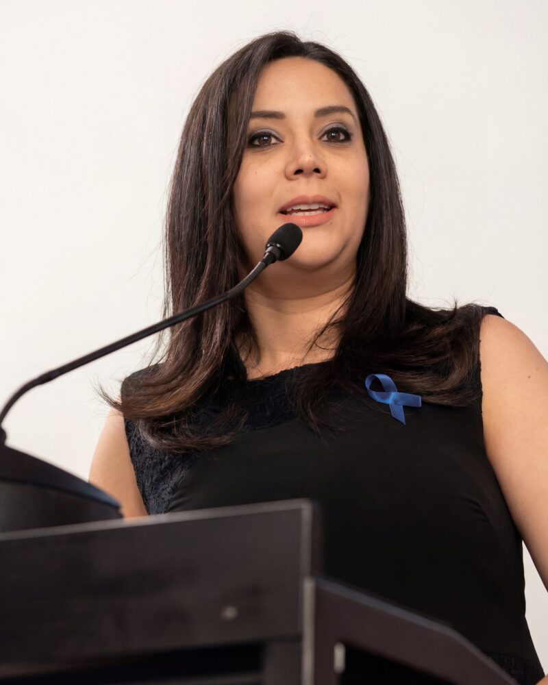 Fotografia colorida. Mulher de cabelos longos e castanhos escuros, com ondas suaves, fala em um microfone. Ela tem uma expressão séria e o olhar focado. Veste uma blusa escura com detalhes de renda e um laço azul vibrante está preso em seu ombro esquerdo.