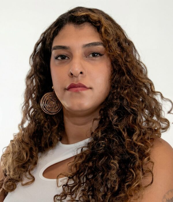 Fotografia colorida. Mulher de cabelos longos, cacheados e volumosos, em tons de castanho com mechas mais claras. Seu rosto oval tem uma expressão séria, com o olhar fixo para a frente. Ela veste uma blusa branca com um recorte no ombro e usa um brinco grande em espiral dourada na orelha direita.