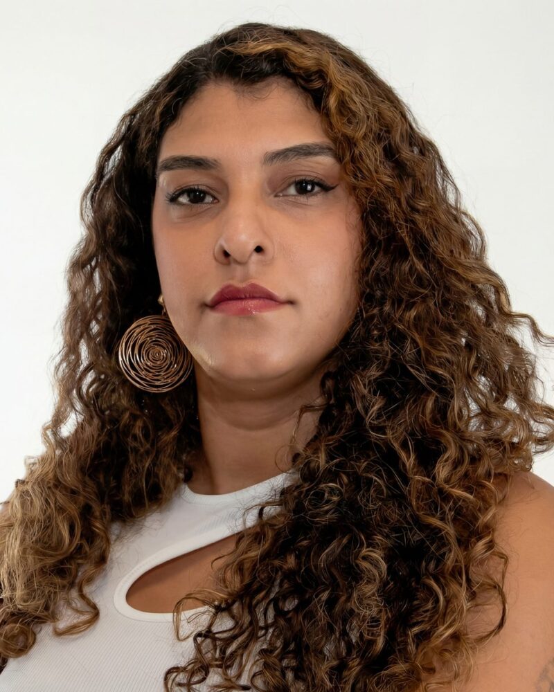 Fotografia colorida. Mulher de cabelos longos, cacheados e volumosos, em tons de castanho com mechas mais claras. Seu rosto oval tem uma expressão séria, com o olhar fixo para a frente. Ela veste uma blusa branca com um recorte no ombro e usa um brinco grande em espiral dourada na orelha direita.