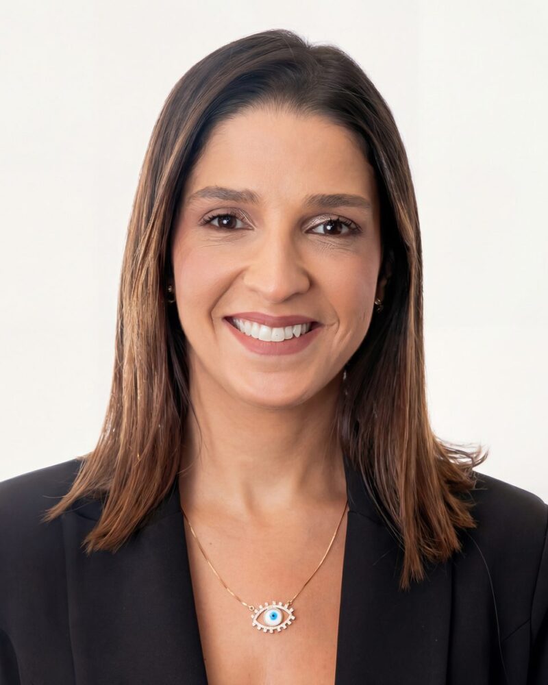 Fotografia colorida. Mulher de cabelos castanhos escuros, lisos e longos, com risca central, caem sobre os ombros. Ela tem um rosto oval e sorri abertamente, olhando diretamente para a frente. Veste um blazer preto e usa um colar fino dourado e brincos pequenos.
