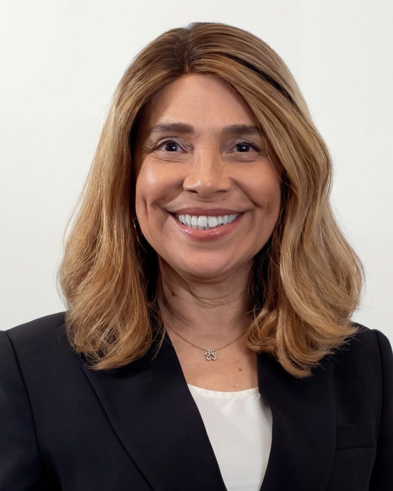 Fotografia colorida. Mulher de cabelos castanhos claros, ondulados e na altura dos ombros, sorri amplamente para a câmera. Ela tem o rosto oval, com covinhas nas bochechas. Veste um blazer escuro sobre uma blusa clara e usa um colar dourado com um pingente de pata.