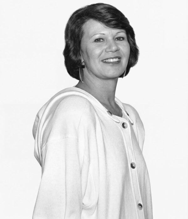 Fotografia em preto e branco. Mulher de cabelos escuros e ondulados, cortados na altura do queixo e com franja lateral. Ela tem um rosto oval, sorri abertamente mostrando os dentes e seu olhar é direto. Veste uma jaqueta clara com botões grandes e usa brincos de argola.