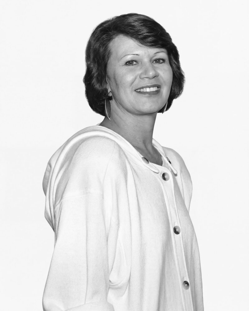 Fotografia em preto e branco. Mulher de cabelos escuros e ondulados, cortados na altura do queixo e com franja lateral. Ela tem um rosto oval, sorri abertamente mostrando os dentes e seu olhar é direto. Veste uma jaqueta clara com botões grandes e usa brincos de argola.