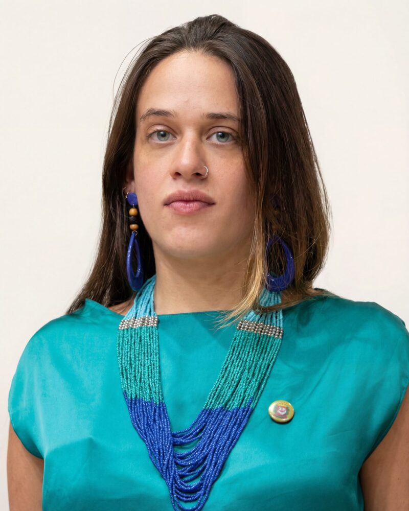 Fotografia colorida. Vereadora branca de cabelos longos e lisos, castanho-escuros, com uma mecha mais clara à esquerda, caindo sobre os ombros. Ela tem um rosto oval, olhos claros e uma expressão neutra, olhando diretamente para a frente. Usa um piercing discreto no nariz, brincos longos azuis e um colar de várias voltas de miçangas turquesa sobre uma blusa da mesma cor.