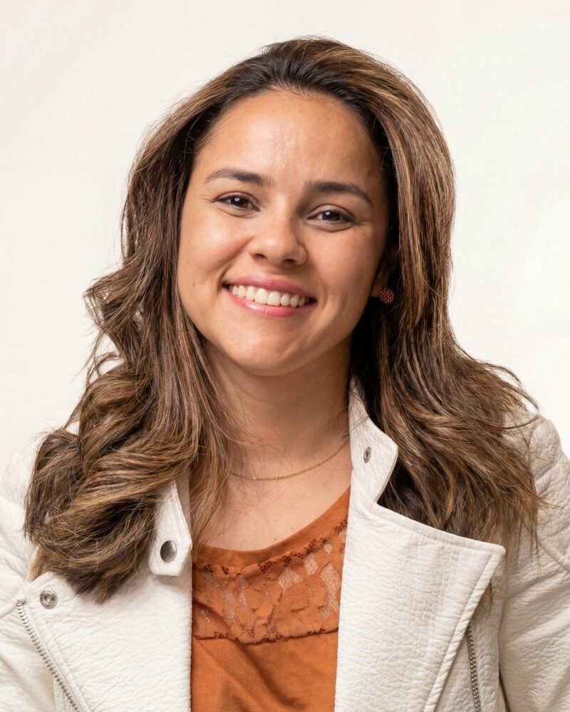 Fotografia colorida. Mulher de cabelos longos e ondulados, castanhos com mechas mais claras, sorri abertamente para a câmera. Ela tem o rosto oval e usa uma jaqueta branca acolchoada sobre uma blusa de renda laranja. Um colar fino dourado e brincos pequenos completam seu visual.