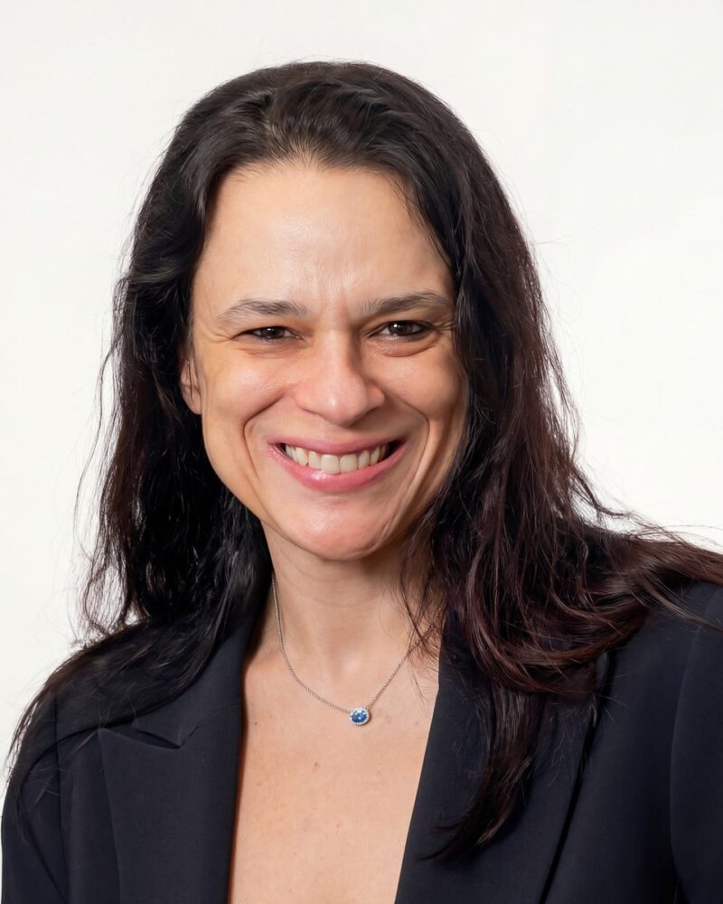 Fotografia colorida. Mulher de cabelos longos e escuros, com ondas suaves, que caem sobre os ombros. Ela tem um rosto oval e sorri abertamente, mostrando os dentes, com um olhar direto para a câmera. Veste um blazer escuro e um colar de corrente fina prateada com um pequeno pingente oval azul, rodeado por pedras brancas.