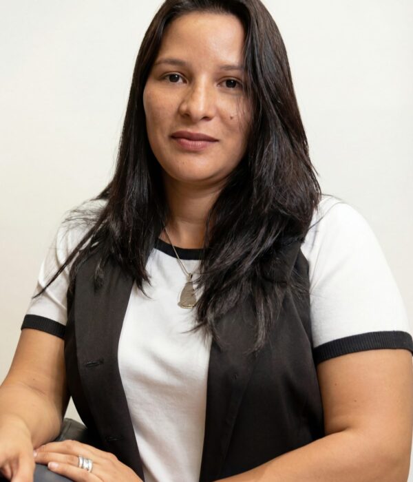 Fotografia colorida. Mulher de cabelos longos e escuros, lisos, caídos sobre os ombros. Ela tem um rosto oval, um leve sorriso e olha diretamente para a câmera. Veste uma camiseta branca com detalhes pretos e um colete escuro, complementado por um colar prateado com um pingente.