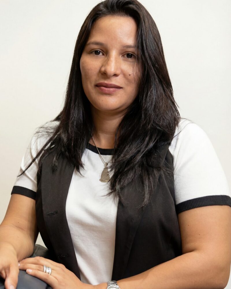 Fotografia colorida. Mulher de cabelos longos e escuros, lisos, caídos sobre os ombros. Ela tem um rosto oval, um leve sorriso e olha diretamente para a câmera. Veste uma camiseta branca com detalhes pretos e um colete escuro, complementado por um colar prateado com um pingente.