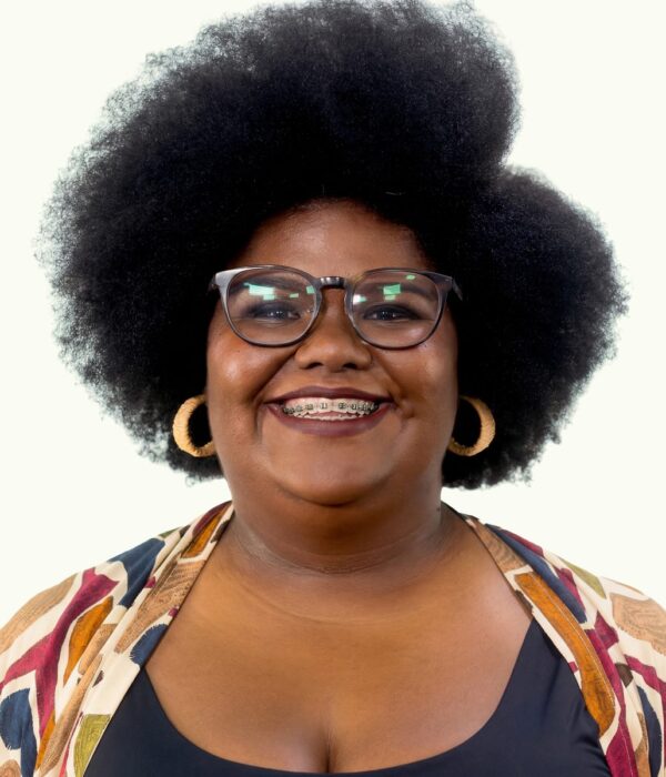Fotografia colorida. Vereadora negra de cabelos pretos, volumosos e crespos, em estilo afro. Ela tem o rosto redondo, sorri abertamente revelando um aparelho ortodôntico e seu olhar é direto e alegre por trás de óculos de aro grosso e escuro. Usa brincos de argola grandes, de textura rústica, e uma blusa colorida com estampa geométrica sobre uma peça escura.
