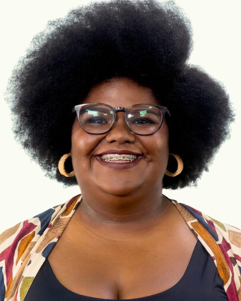 Fotografia colorida. Vereadora negra de cabelos pretos, volumosos e crespos, em estilo afro. Ela tem o rosto redondo, sorri abertamente revelando um aparelho ortodôntico e seu olhar é direto e alegre por trás de óculos de aro grosso e escuro. Usa brincos de argola grandes, de textura rústica, e uma blusa colorida com estampa geométrica sobre uma peça escura.