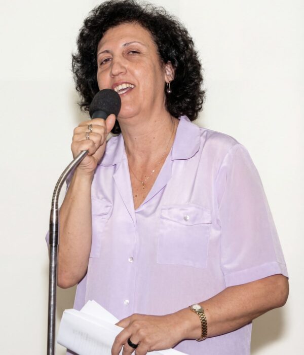 Fotografia colorida. Mulher de cabelos escuros e encaracolados, com rosto oval, sorri abertamente enquanto segura um microfone com a mão direita e está falando. Ela usa uma camisa de botão de cor clara com bolsos no peito, um colar fino com pingente e um anel na mão direita. Seus olhos estão ligeiramente semicerrados.