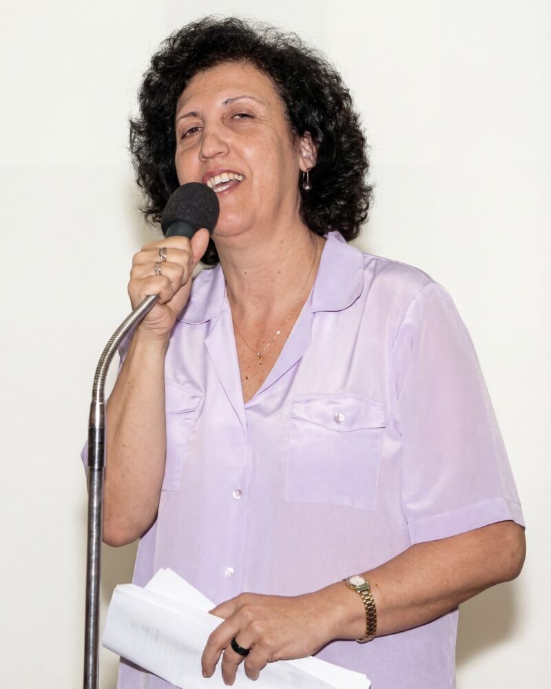 Fotografia colorida. Mulher de cabelos escuros e encaracolados, com rosto oval, sorri abertamente enquanto segura um microfone com a mão direita e está falando. Ela usa uma camisa de botão de cor clara com bolsos no peito, um colar fino com pingente e um anel na mão direita. Seus olhos estão ligeiramente semicerrados.