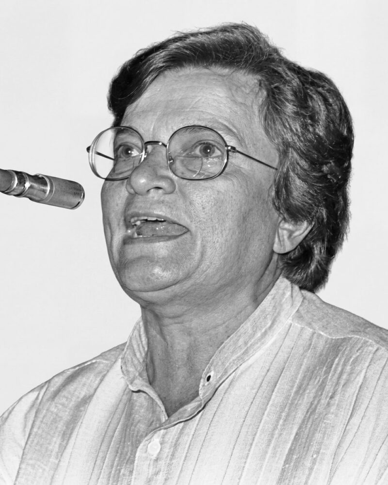 Fotografia em preto e branco. Vereadora branca de cabelos ondulados e claros, penteados para trás. Ela usa óculos redondos de aro fino e tem uma expressão intensa, com a boca aberta como se estivesse falando e o olhar direcionado para cima. Veste uma camisa listrada de colarinho.
