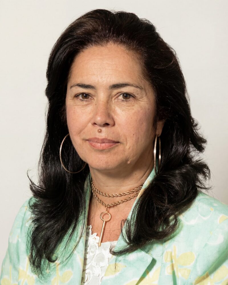 Fotografia colorida. Mulher de cabelos longos e ondulados, de cor escura, que caem sobre os ombros. Ela tem um rosto oval, sobrancelhas cheias e um olhar direto e calmo. Veste uma blusa clara e usa brincos de argola grandes e dois colares de corrente dourados.