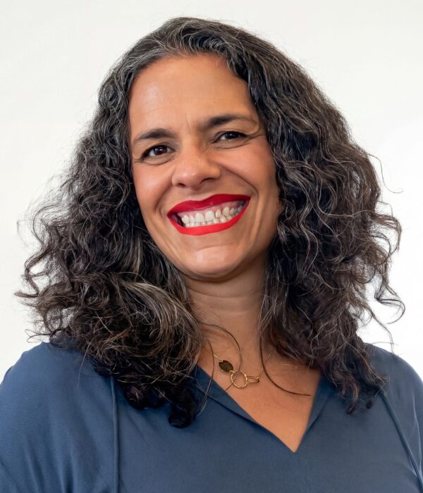 Fotografia colorida. Mulher de cabelos longos, escuros e cacheados, com fios grisalhos visíveis. Ela tem um rosto oval e sorri abertamente, mostrando os dentes, com um olhar direto. Veste uma blusa azul-marinho com decote em V e usa um colar dourado com elos circulares.