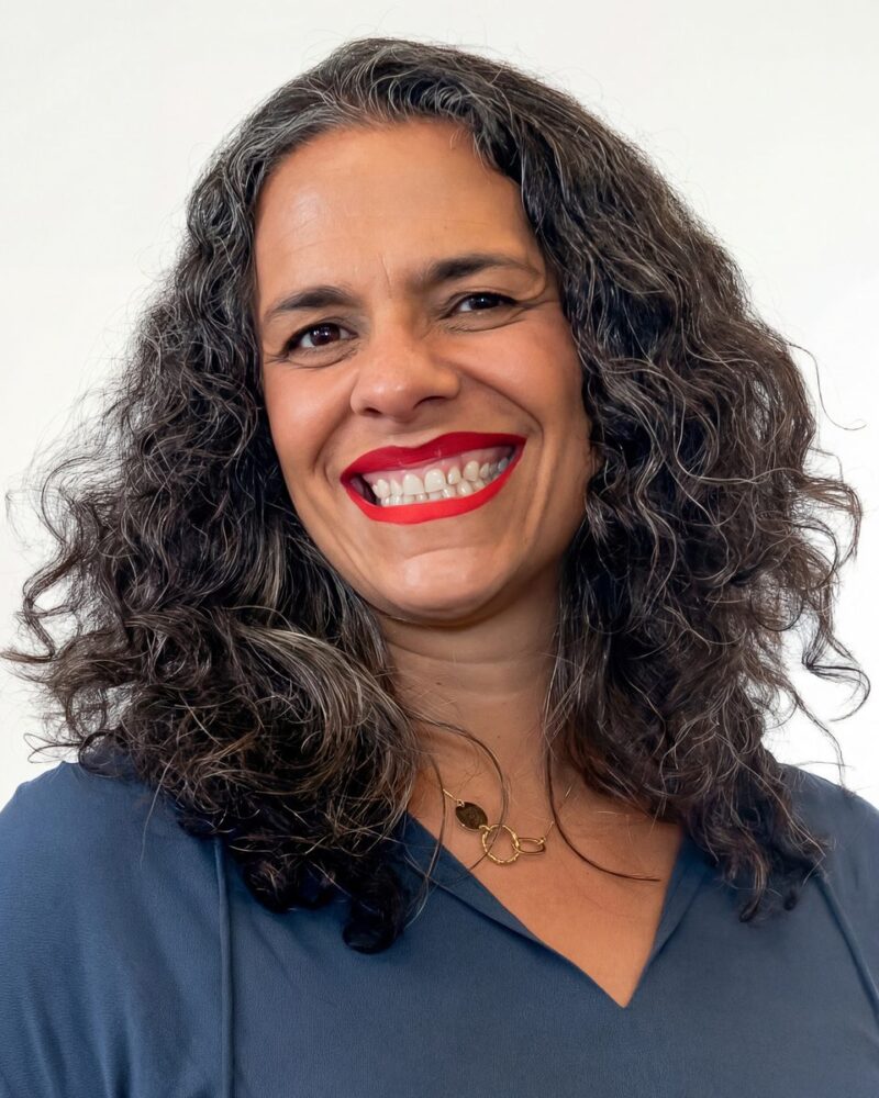 Fotografia colorida. Mulher de cabelos longos, escuros e cacheados, com fios grisalhos visíveis. Ela tem um rosto oval e sorri abertamente, mostrando os dentes, com um olhar direto. Veste uma blusa azul-marinho com decote em V e usa um colar dourado com elos circulares.