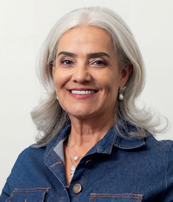 Fotografia colorida. Vereadora branca de cabelos grisalhos ondulados na altura dos ombros. Ela sorri abertamente, mostrando os dentes, e olha diretamente para a câmera. Veste uma camisa jeans azul com costuras laranja e usa um colar prateado com pingentes, além de brincos de borboleta com pérolas.