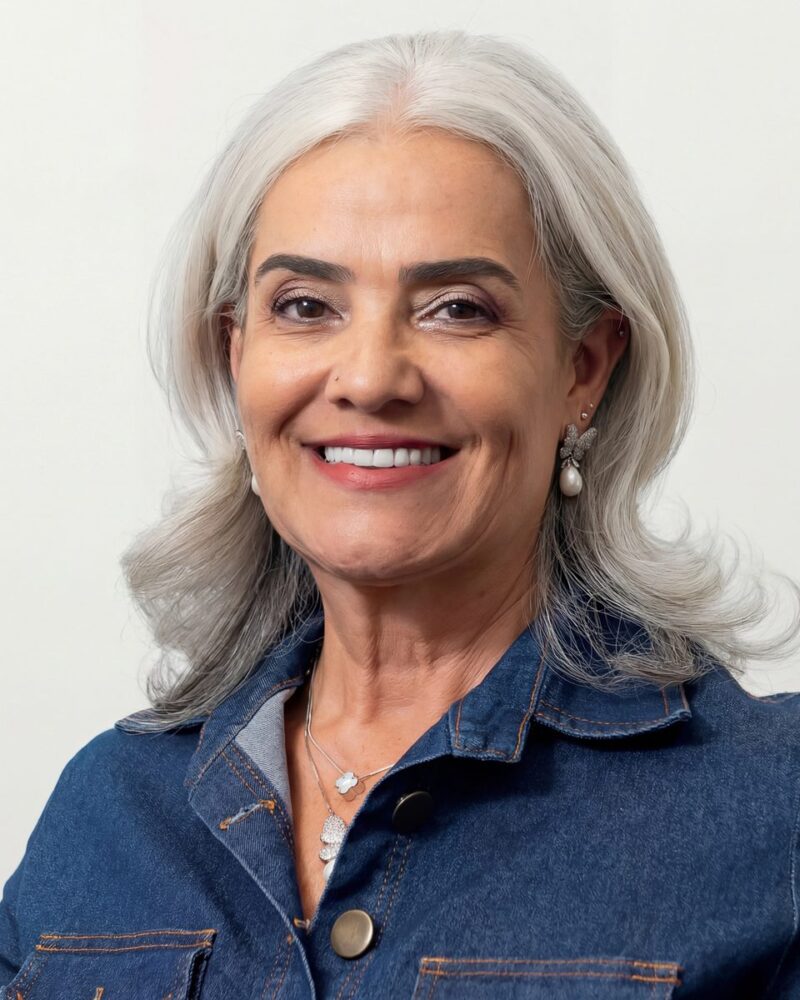 Fotografia colorida. Vereadora branca de cabelos grisalhos ondulados na altura dos ombros. Ela sorri abertamente, mostrando os dentes, e olha diretamente para a câmera. Veste uma camisa jeans azul com costuras laranja e usa um colar prateado com pingentes, além de brincos de borboleta com pérolas.