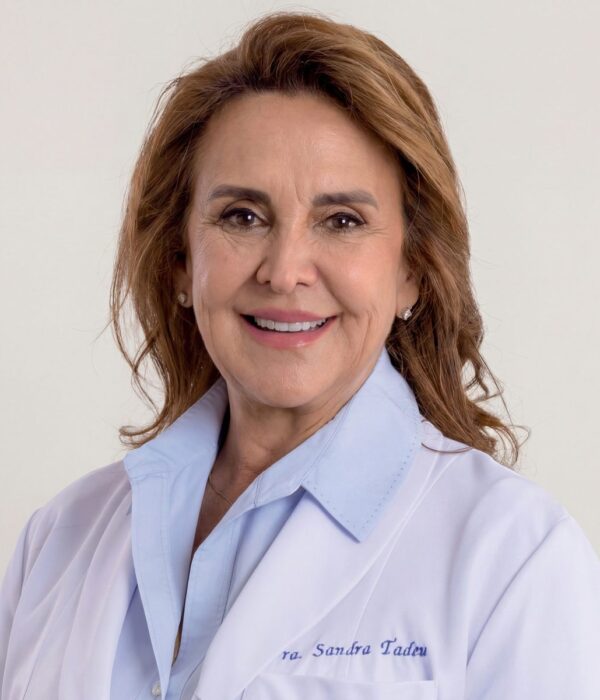 Fotografia colorida. Vereadora branca de cabelos castanhos claros, ondulados e na altura dos ombros. Ela sorri amplamente, mostrando os dentes, e tem um olhar direto para a frente. Veste uma camisa azul clara com colarinho e um casaco branco, usando brincos de ponto de luz e um colar fino.