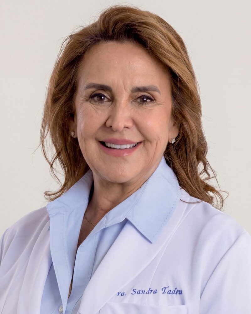Fotografia colorida. Vereadora branca de cabelos castanhos claros, ondulados e na altura dos ombros. Ela sorri amplamente, mostrando os dentes, e tem um olhar direto para a frente. Veste uma camisa azul clara com colarinho e um casaco branco, usando brincos de ponto de luz e um colar fino.