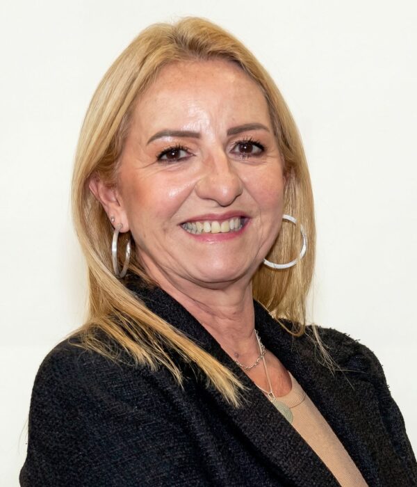 Fotografia colorida. Vereadora branca de cabelos loiros e lisos na altura dos ombros. Ela sorri abertamente, mostrando os dentes, e usa grandes brincos de argola prateados. Veste um blazer escuro sobre uma blusa clara e um colar prateado.