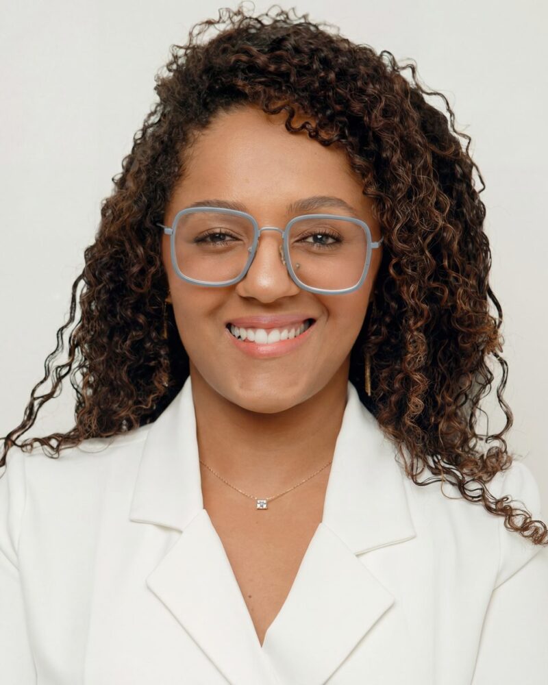 Fotografia colorida. Vereadora negra de cabelos longos, escuros e cacheados, soltos sobre os ombros. Ela sorri abertamente, mostrando os dentes, e usa óculos grandes de aro quadrado azul-claro. Veste um blazer branco e um colar fino dourado com um pequeno pingente quadrado.