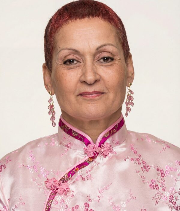 Fotografia colorida. Mulher de cabelos curtos e avermelhados, com um rosto oval e expressão serena. Ela usa uma blusa rosa clara com gola alta e detalhes em rosa escuro bordados. Brincos longos com pedras rosadas adornam suas orelhas.