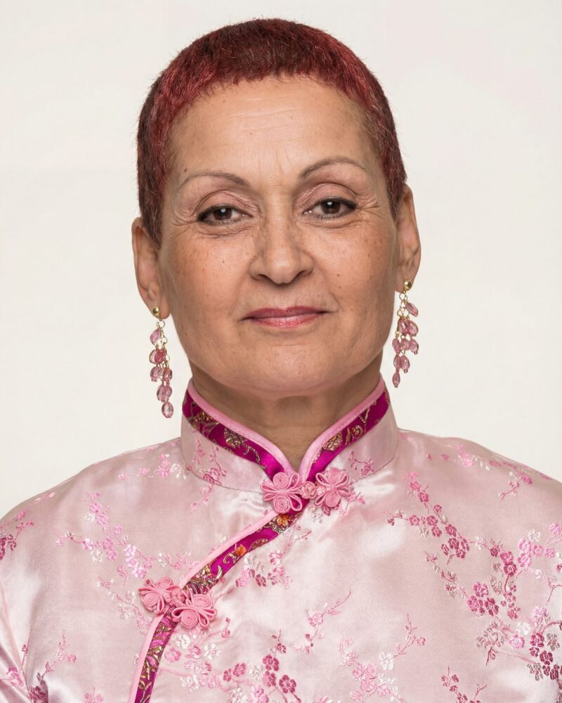 Fotografia colorida. Mulher de cabelos curtos e avermelhados, com um rosto oval e expressão serena. Ela usa uma blusa rosa clara com gola alta e detalhes em rosa escuro bordados. Brincos longos com pedras rosadas adornam suas orelhas.