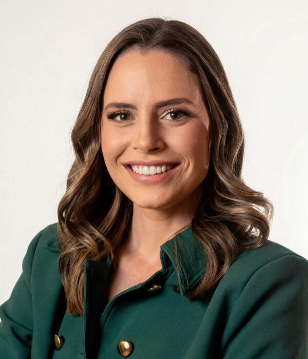Fotografia colorida. Vereadora branca de cabelos longos e ondulados, castanhos com mechas claras, que caem sobre os ombros. Ela tem um rosto oval e sorri abertamente, olhando diretamente para a câmera. Veste um blazer verde-escuro com botões dourados.