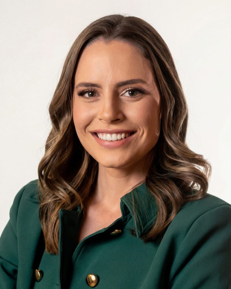 Fotografia colorida. Vereadora branca de cabelos longos e ondulados, castanhos com mechas claras, que caem sobre os ombros. Ela tem um rosto oval e sorri abertamente, olhando diretamente para a câmera. Veste um blazer verde-escuro com botões dourados.