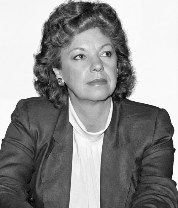 Fotografia em preto e branco. Vereadora branca de cabelos escuros ondulados e volumosos, na altura dos ombros. Ela tem um rosto oval e uma expressão séria, com o olhar direcionado para cima e para a direita. Veste um blazer escuro sobre uma blusa clara de gola alta e usa brincos ornamentados.