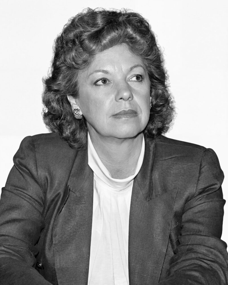 Fotografia em preto e branco. Vereadora branca de cabelos escuros ondulados e volumosos, na altura dos ombros. Ela tem um rosto oval e uma expressão séria, com o olhar direcionado para cima e para a direita. Veste um blazer escuro sobre uma blusa clara de gola alta e usa brincos ornamentados.